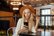 Indoor photo of pretty young long haired blonde female sitting at table over cafe interior, keeping smartphone in hand and looking at screen with soft smile