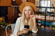 Young attractive blonde woman with long hair sitting at cafe table and looking dreamily ahead, touching her chi with and and holding smartphone