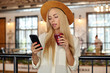 Young pretty blonde female in white shirt and wide hat drinking lemonade while lunch break, holding mobile phone in hand and looking at screen with calm face