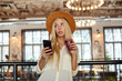 Attractive young blonde female drinking smoothie with straw while waiting for her order in cafe, holding smartphone in hand and looking aside with interest