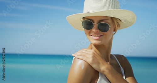 A happy young blond hair woman with a hat and sunglasses is applying a sunscreen or sun tanning lotion to take care of her skin during a vacation on a beach.