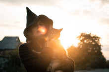 Halloween Cat In Hands And A Witch Girl Wearing A Black Hat And Coat On An Autumn Sunset. Celebration And Pets Concept.