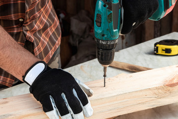 A man carpenter twists a screw into a tree with an electric screwdriver, male hands with a screwdriver close-up. Work with wood.