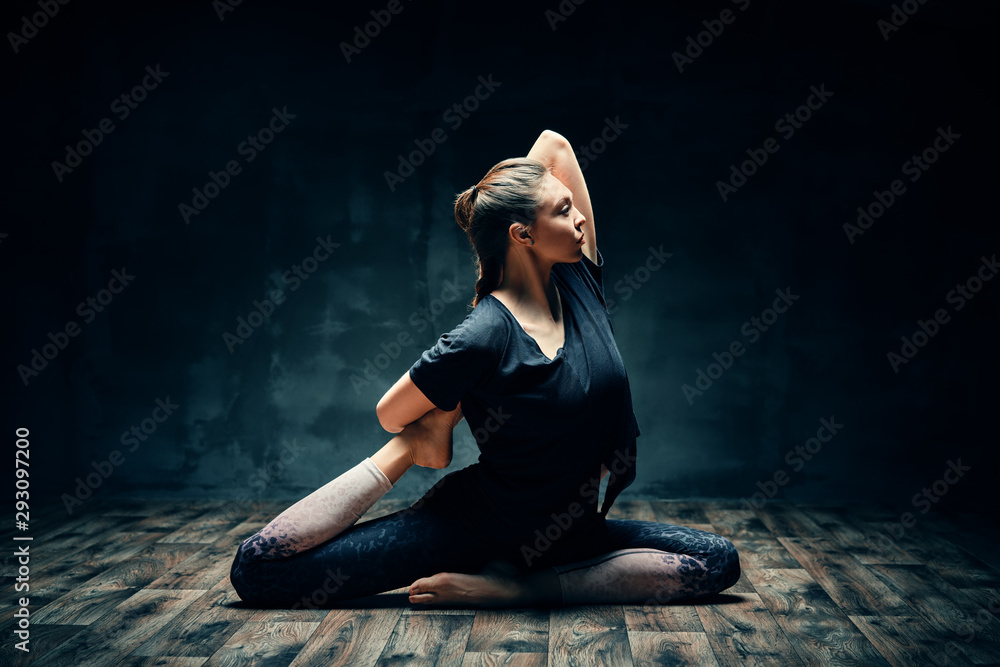 Young woman practicing yoga doing one legged king pigeon pose in dark room - obrazy, fototapety, plakaty 