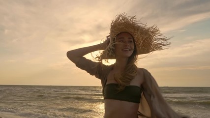 Wall Mural - Cheerful pretty young woman holding her straw hat and relaxing while running at the seaside