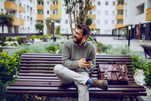 Laughing Handsome Stylish Businessman Typing Message On Smart Phone While Sitting On The Bench In Park. Next To Him Is Leather Bag.