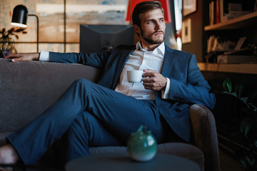 Wall Mural - Handsome young man holding coffee cup sitting on the couch in office