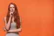 © timtimphoto - Studio shot of young attractive red haired female in casual clothes standing over orange background, holding chin with hand and looking aside pensively, trying to gather with thoughts