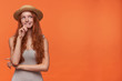 Lovely young wavy lond haired foxy woman in grey shirt and straw hat holding her chin, looking aside with dreamy face and biting underlip, isolated over orange background