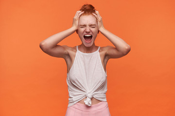 Wall Mural - Studio close-up of stressed young woman with foxy hair combed in bun standin over orange background, keeping hands on temples and screaming loud with closed eyes
