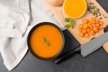 Wall Mural - food, new nordic cuisine, culinary and cooking concept - close up of pumpkin cream soup in bowl, chopped vegetables and knife on stone table