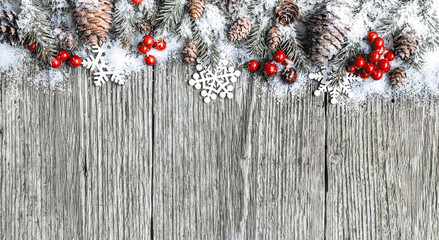 Christmas frame made of fir branches, cones and red christmas berries covered by snow on wood desk.