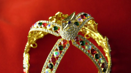 Gold diamond crown or decorative pageant accessory close up focus on the top. On the red royal color surface.