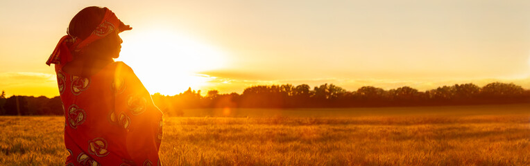 African woman in traditional clothes in field of crops at sunset or sunrise panorama