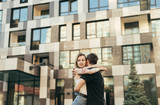 Fototapeta Na drzwi - Beautiful young couple hugging on the street on the background of a modern building, the girl looks away. Guy hugged the girl on a walk on a warm summer day, she is happy. Love story