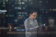 © RolandoMayo - Side view shot of a young man in long sleeves and eye glasses standing and reading a book inside an office building at night. View through window glass with blurred building lights reflected.