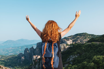 A relaxed red-haired woman with a tourist backpack is standing on top of a high mountain against the backdrop of the Meteora Mountains in Greece, looking at the mountains raising their hands up