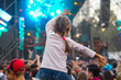 Child has fun on her parents' shoulders keeping hands with them at an outdoor rock music concert