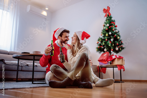 Fotomural Happy handsome caucasian couple with santa hats on heads sitting on the floor with beer bottle in hands and cuddling