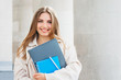 © Evgenia - Young blonde girl student smiling against university. Cute girl student holds folders and notebooks in hands, copy space. Learning, education concept