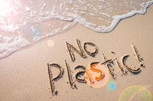 No Plastic! Pollution Awareness Message Handwritten In Smooth Clean Sand With Lens Flare Above An Incoming Wave On A Sunny Beach