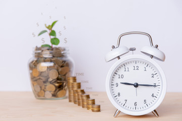 Savings on white background. Concept with white clock, jar and stack of money. This shows that savings are accumulating gradually and slowly. All on a wooden table and a white background.  