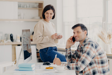 Horizontal shot of cheerful man has telephone conversation, discusses financial contract agreement, dressed in checkered shirt, writes down information, happy woman holds mug of drink, leans at table