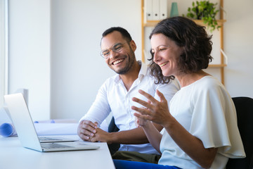 Cheerful colleagues using laptop for video call. Man and woman sitting at workplace together, looking at monitor and laughing. Communication concept