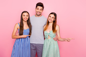 Poster - Portrait of cheerful man and two woman with long ginger foxy hair pointing at copy space wearing striped dress skirt t-shirt isolated over pink background