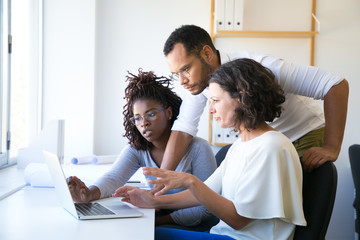 Instructor helping new employees with corporate software. Diverse man and women sitting and standing at desk, using computer, talking. Training concept