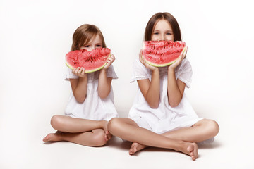 Two little girls, sisters, sit in the studio on a white background and hide behind pieces of watermelon. In bright clothes, barefoot. Happy childhood, pampering. Fruit, proper nutrition.