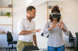© Mangostar - Young woman in VR headset standing in office, touching air. Her male colleague instructing her, showing hand gestures. Virtual reality simulation concept