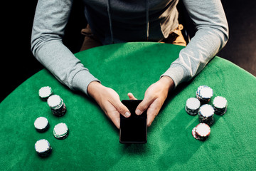 cropped view of man holding smartphone with blank screen near poker chips on black