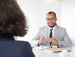 © Mangostar - Smiling businessman talking with female colleague. Young African American businessman in eyeglasses sitting at table with colleague during business meeting. Job interview concept
