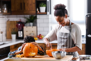 Wall Mural - Young woman with her little son are preparing for Halloween on kitchen. Mother with son are having fun with pumpkin.