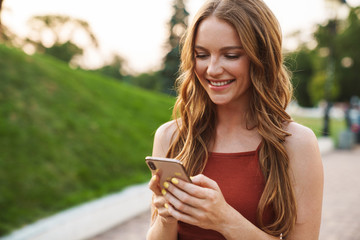 Wall Mural - Ginger woman walking in park outdoors using mobile phone.