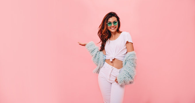 Laughing brunette woman in white t-shirt and mint coat looking and pointing away with thumb over pink background. funny surprised girl showing empty copy space with her hand