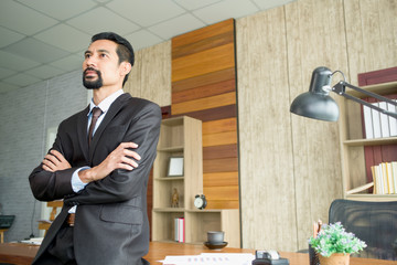 Adult professional businessman wearing the black suit is standing and arms crossed within a modern office.