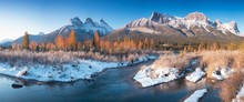Panorama Of Autumn Sunrise At The Three Sisters Mountain With Colorful Trees Canmore, Alberta With Reflection In Calm Water Of Policeman Creek Surrounded By Trees And Bushes. First Snow In Mountains