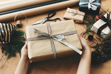 hands holding stylish christmas gift box in craft paper and scissors, rustic presents, thread, pine 