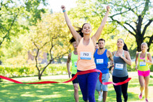 Fitness, Sport And Success Concept - Happy Woman Winning Race And Coming First To Finish Red Ribbon Over Group Of Sportsmen Running Marathon With Badge Numbers At Park