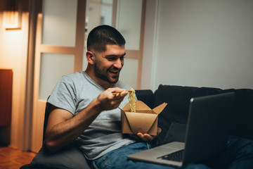 man eating chinese food and using laptop computer relaxed on sofa at his home