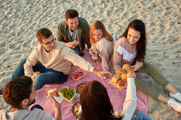 Wall Mural - friendship, leisure and fast food concept - group of happy friends eating sandwiches or burgers at picnic on beach in summer