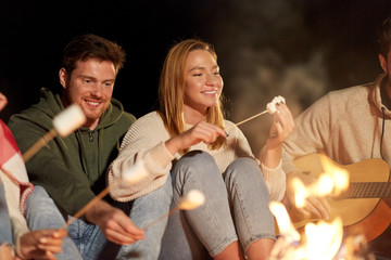 Poster - leisure and people concept - group of smiling friends sitting at camp fire, roasting marshmallow and playing guitar at night