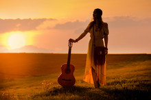 Siluette Of Woman Wearing A Bohemian Style Holding A Guitar On A Field At Warm Light Of Sunset