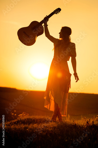 Fototapety Country & Western  sylwetka-kobiety-w-stylu-bohemy-trzymajacej-gitare-na-polu-w-cieplym-swietle-su