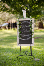 Decorated Welcome To Our Wedding Sign In Grassy Field On A Sunny Day With Ceremony Guests In Background