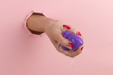 Sticker - Girl hand holds a beauty blender for makeup from a hole in a pink background, copy space. Sponge in soapy foam, how to clean.