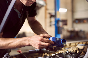 Engine oil changing in auto workshop. Focus on pouring grease into engine from bottle by repairman hands. Cropped view. Blurred background. Maintenance car engine. Automotive service for safe driving