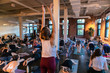 © Valmedia - Diverse group of people in yoga class. A young woman is seen in an upward arm stretch pose during a workshop dedicated to 108 sun salutes. Standing alone as others are seen in downward postures.